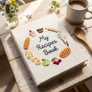 Hand embroidered recipe book featuring cupcakes, pie, artisan bread, pasta, fruit and baking tools on linen cover.