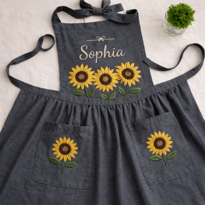 Personalized hand embroidered linen apron featuring three sunflower blooms, botanical foliage, floral pocket embroidery, and custom name personalization.