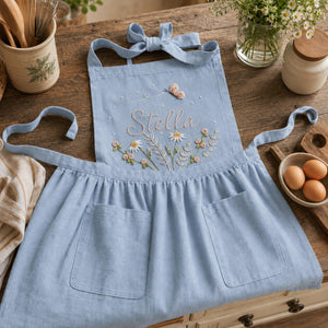 Personalized hand embroidered linen apron featuring wildflowers, daisies, botanical foliage, butterfly embroidery, and custom name embroidery on green linen fabric.