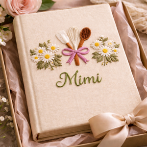 Personalized hand embroidered recipe book featuring a whisk, spatula, wooden spoon tied with a pink ribbon and surrounded by white daisies on a linen cover.