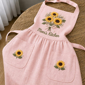 Personalized hand embroidered linen apron featuring a sunflower bouquet, white floral accents, ribbon detail, and custom name embroidery.