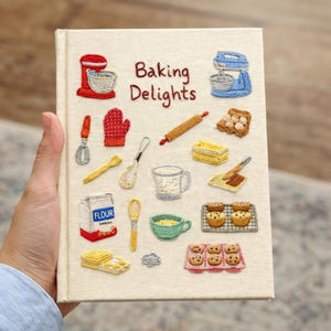Hand embroidered recipe book featuring vintage baking studio essentials including stand mixers, rolling pins, flour, eggs, whisks, muffins and cookies on a linen cover.
