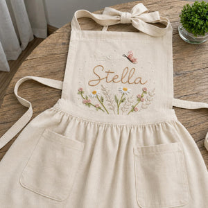 Personalized hand embroidered linen apron featuring wildflowers, daisies, botanical foliage, butterfly embroidery, and custom name embroidery on green linen fabric.