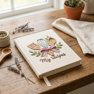 Hand embroidered recipe book featuring whisk, wooden spoon, spatula, pink ribbon and leafy branches on linen cover.