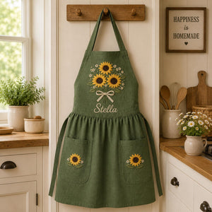 Personalized hand embroidered linen apron featuring a sunflower bouquet, white floral accents, ribbon detail, and custom name embroidery.