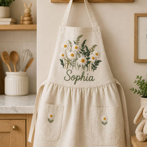 Personalized hand embroidered daisy flower apron featuring custom name embroidery, white daisies, and botanical greenery on natural linen fabric.