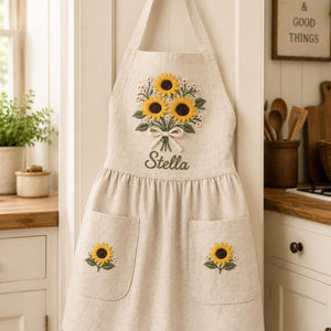 Personalized hand embroidered linen apron featuring a sunflower bouquet, white floral accents, ribbon detail, and custom name embroidery.