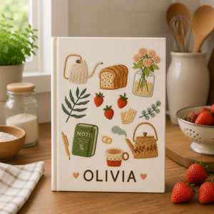 Personalized hand embroidered recipe book featuring cottage kitchen motifs, strawberries, teapot, herbs, lemons, avocado, olive oil and fresh cooking ingredients on linen cover.