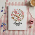 Personalized hand embroidered recipe book featuring pink floral wreath with rolling pin, whisk and wooden spoon on a white linen cover.