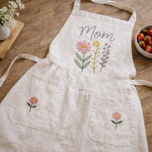 Personalized hand embroidered linen apron featuring pink wildflowers, botanical floral bouquet embroidery, custom name personalization, and floral pocket details.