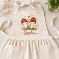 Personalized hand embroidered apron featuring red toadstool mushrooms, daisy flowers, meadow grass, and custom name embroidery on natural linen fabric.