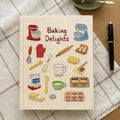 Hand embroidered recipe book featuring vintage baking studio essentials including stand mixers, rolling pins, flour, eggs, whisks, muffins and cookies on a linen cover.