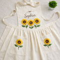 Personalized hand embroidered linen apron featuring three sunflower blooms, botanical foliage, floral pocket embroidery, and custom name personalization.