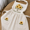 Personalized hand embroidered linen apron featuring a sunflower bouquet, white floral accents, ribbon detail, and custom name embroidery.