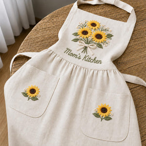 Personalized hand embroidered linen apron featuring a sunflower bouquet, white floral accents, ribbon detail, and custom name embroidery.