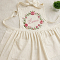 Personalized hand embroidered apron featuring floral wreath embroidery with custom name surrounded by pink roses and botanical accents.