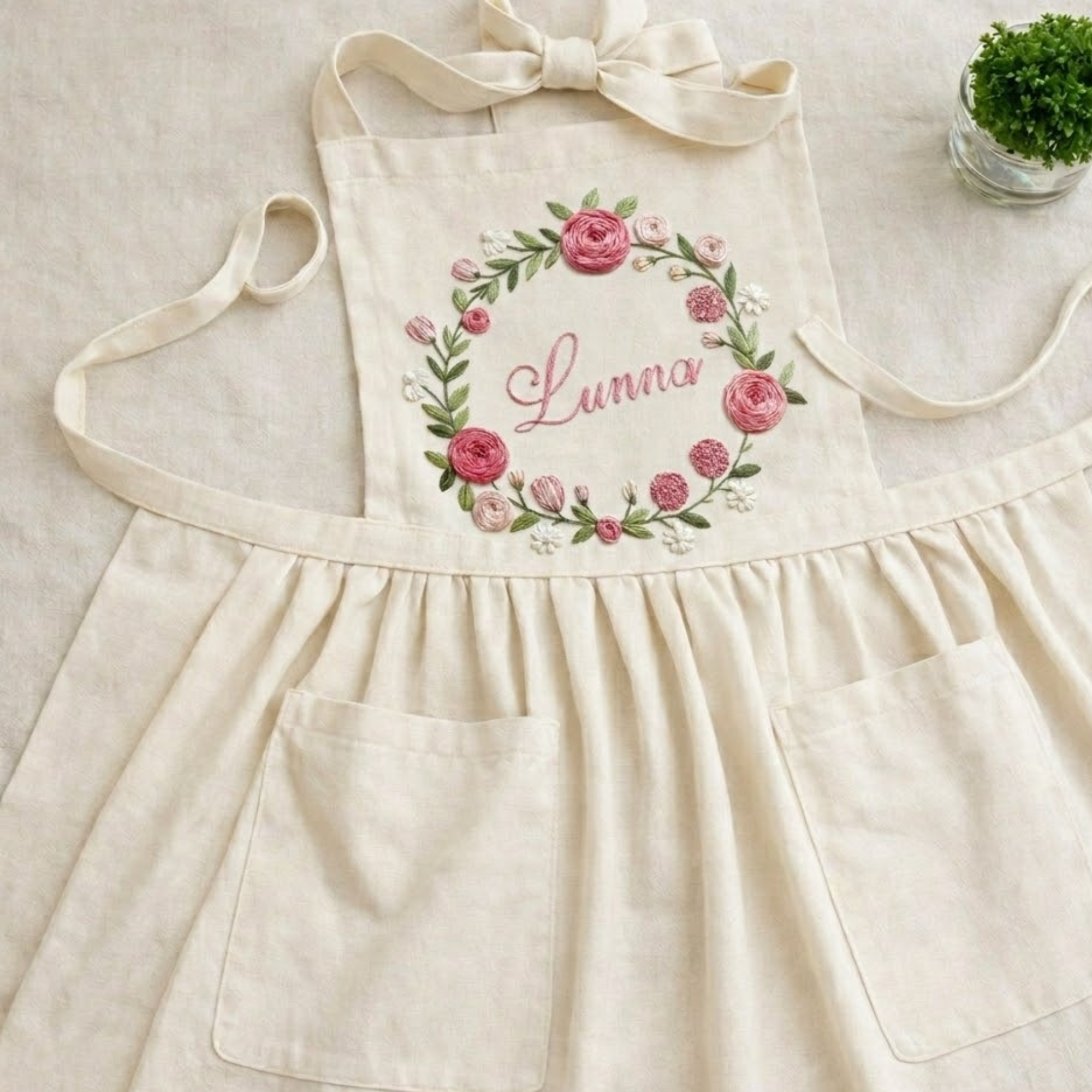 Personalized hand embroidered apron featuring floral wreath embroidery with custom name surrounded by pink roses and botanical accents.