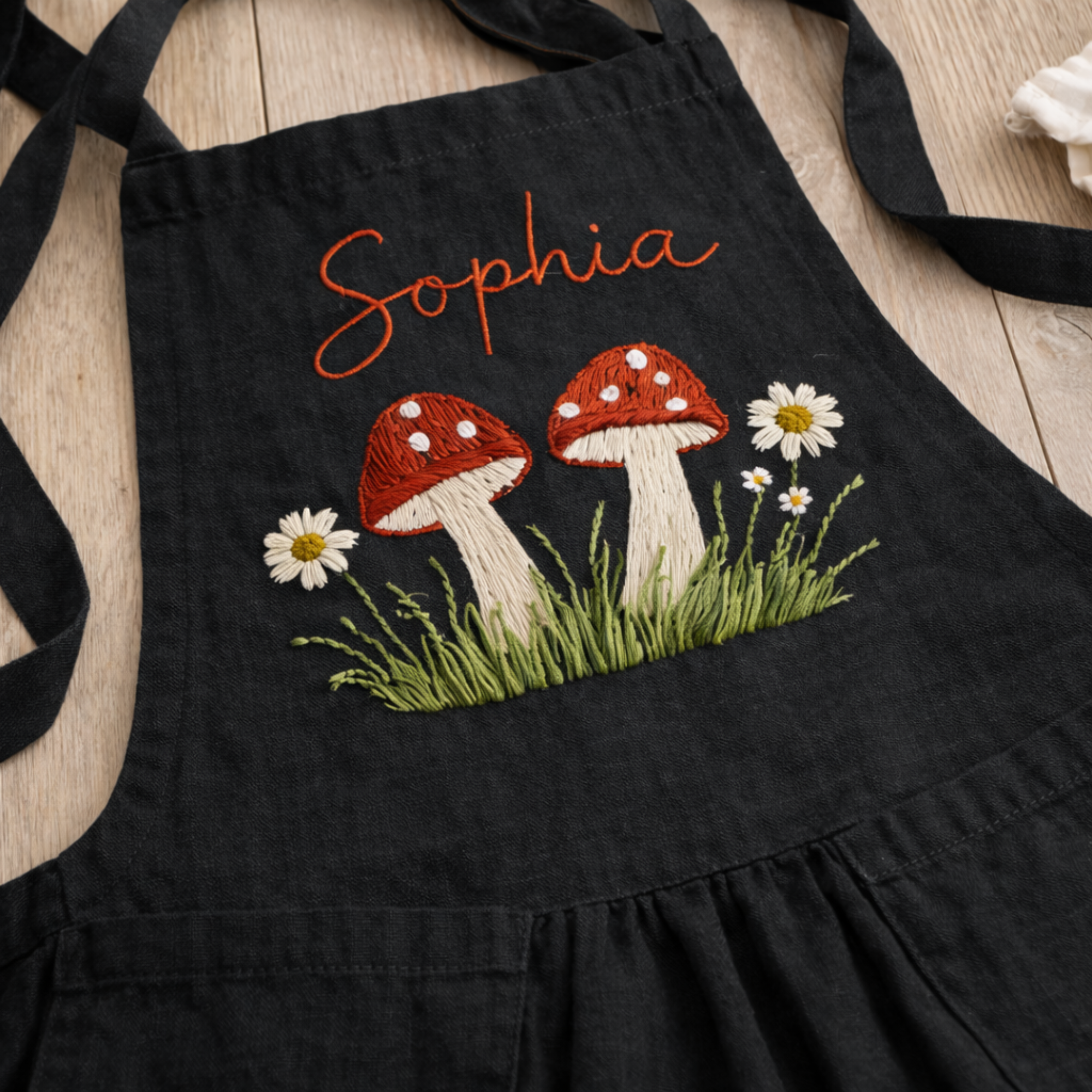 Personalized hand embroidered linen apron featuring red woodland mushrooms, wild daisies, green meadow grass and custom name embroidery in cottagecore style.