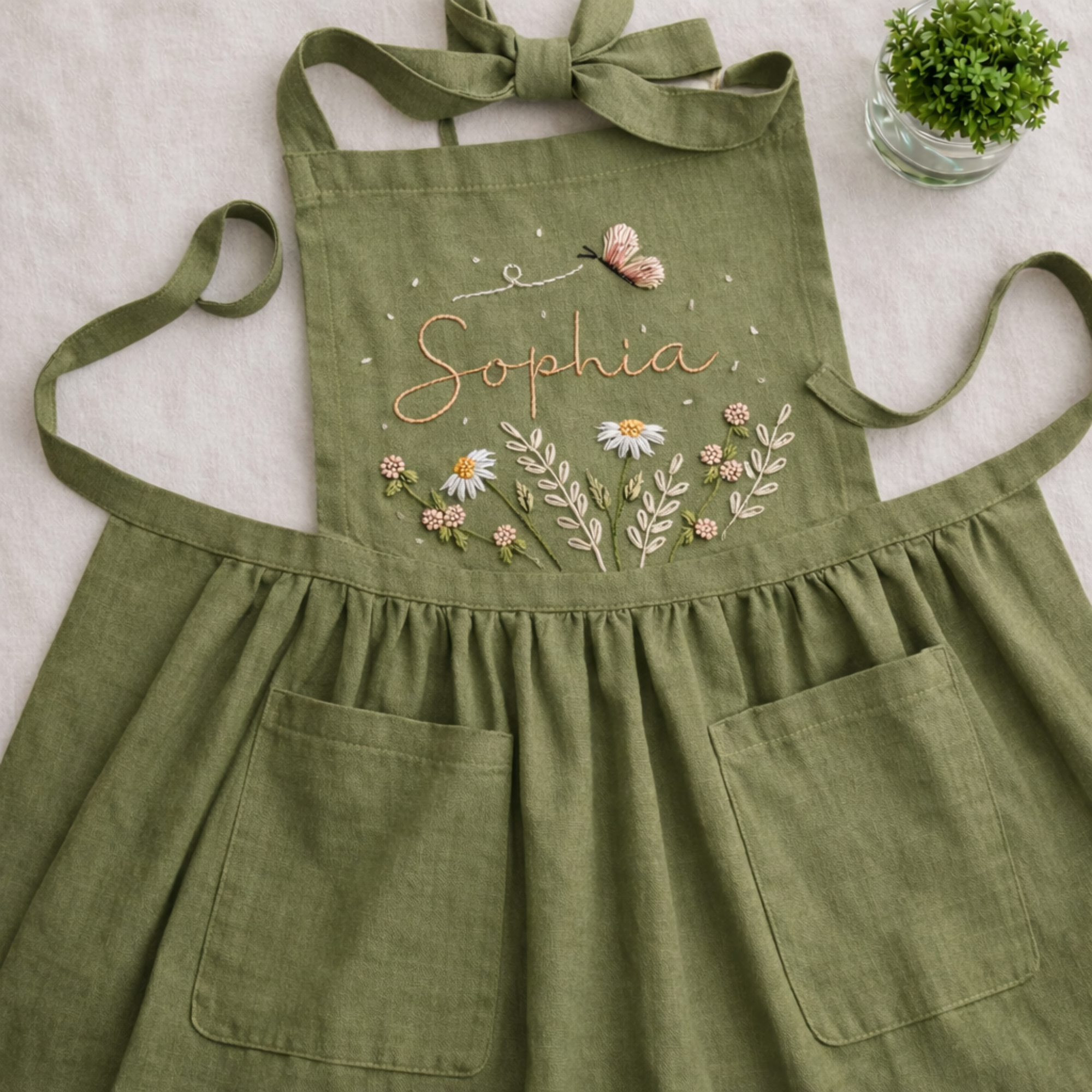 Personalized hand embroidered linen apron featuring wildflowers, daisy flowers, botanical foliage, butterfly embroidery, and custom name personalization on green linen fabric.