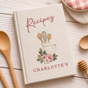 Hand embroidered recipe book featuring a vintage utensil pitcher with pink roses and personalized name on a linen cover.