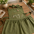 Personalized hand embroidered linen apron featuring wildflowers, daisies, botanical foliage, butterfly embroidery, and custom name embroidery on green linen fabric.