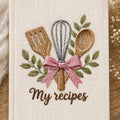 Hand embroidered recipe book featuring whisk, wooden spoon, spatula, pink ribbon and leafy branches on linen cover.