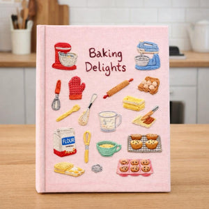 Hand embroidered recipe book featuring vintage baking studio essentials including stand mixers, rolling pins, flour, eggs, whisks, muffins and cookies on a linen cover.