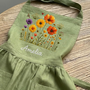 Personalized hand embroidered linen apron featuring colorful wildflowers, meadow flowers, botanical embroidery, and custom name embroidery on green linen fabric.