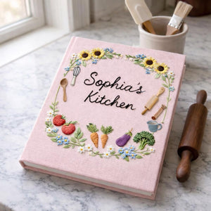 Personalized hand embroidered recipe book featuring sunflowers, strawberries, carrots, broccoli, baking tools and cottage garden floral wreath design on linen cover.