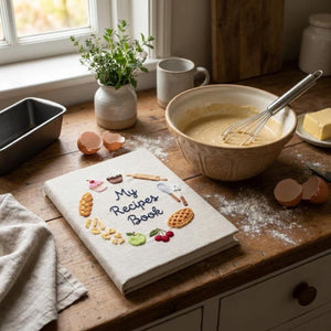 Hand embroidered recipe book featuring cupcakes, pie, artisan bread, pasta, fruit and baking tools on linen cover.