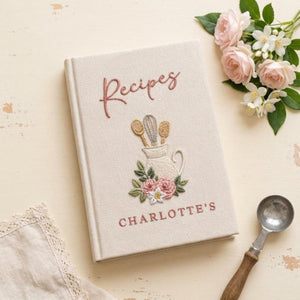 Hand embroidered recipe book featuring a vintage utensil pitcher with pink roses and personalized name on a linen cover.