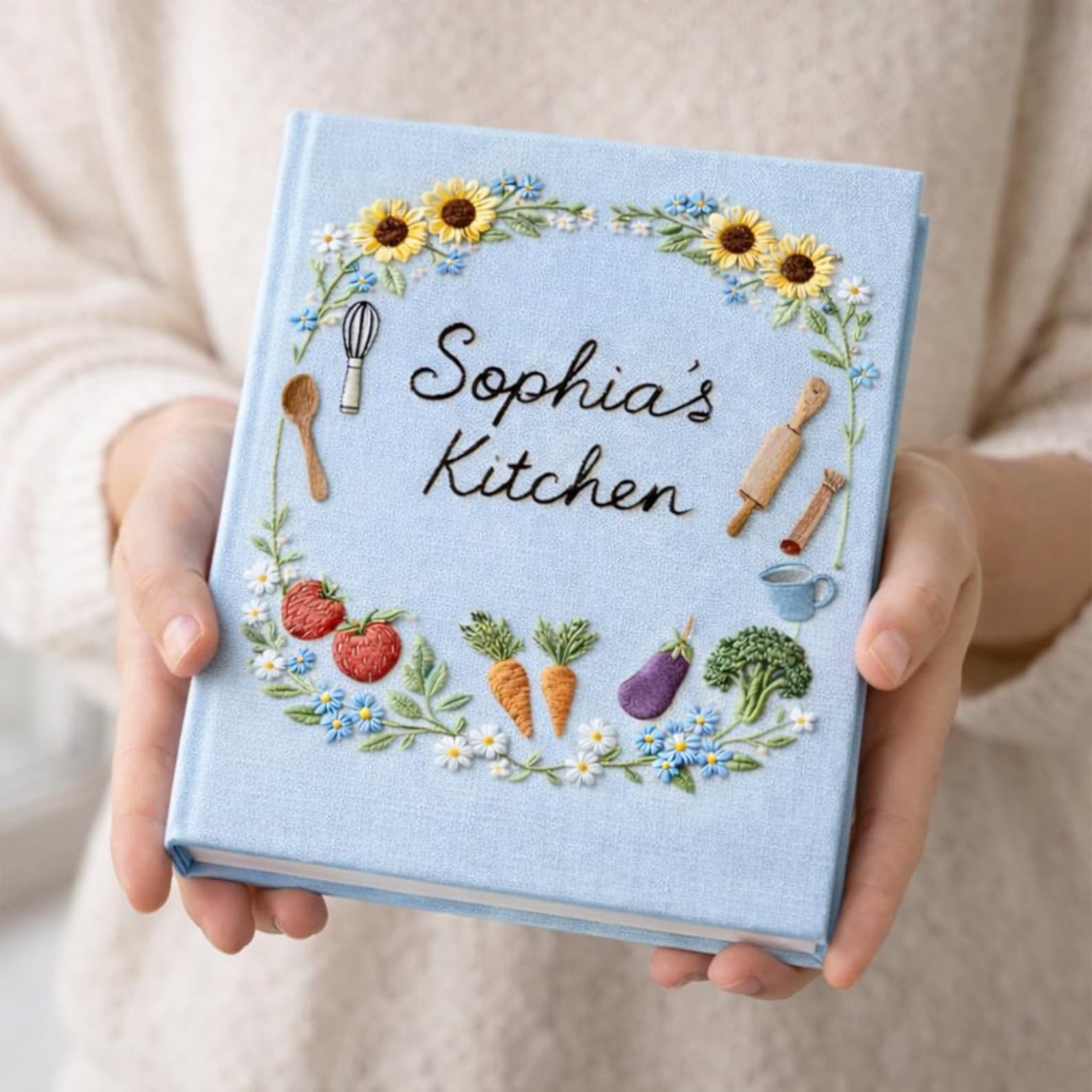 Personalized hand embroidered recipe book featuring sunflowers, strawberries, carrots, broccoli, baking tools and cottage garden floral wreath design on linen cover.