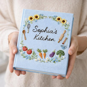 Personalized hand embroidered recipe book featuring sunflowers, strawberries, carrots, broccoli, baking tools and cottage garden floral wreath design on linen cover.