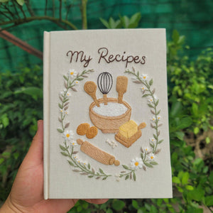 Hand embroidered recipe book featuring mixing bowl flour eggs butter rolling pin whisk and daisy floral wreath on linen cover