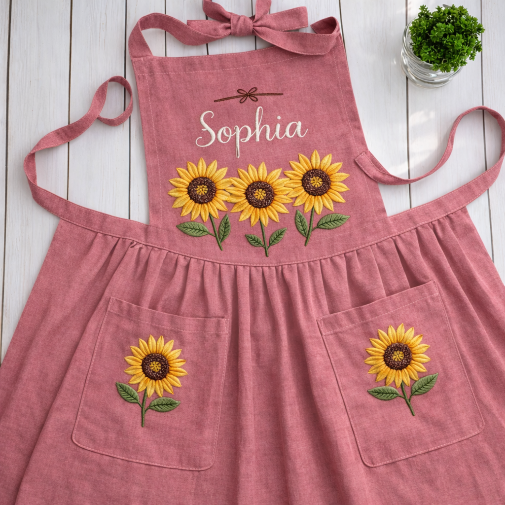 Personalized hand embroidered linen apron featuring three sunflower blooms, botanical foliage, floral pocket embroidery, and custom name personalization.
