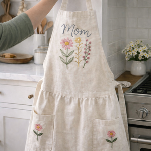 Personalized hand embroidered linen apron featuring pink wildflowers, botanical floral bouquet embroidery, custom name personalization, and floral pocket details.