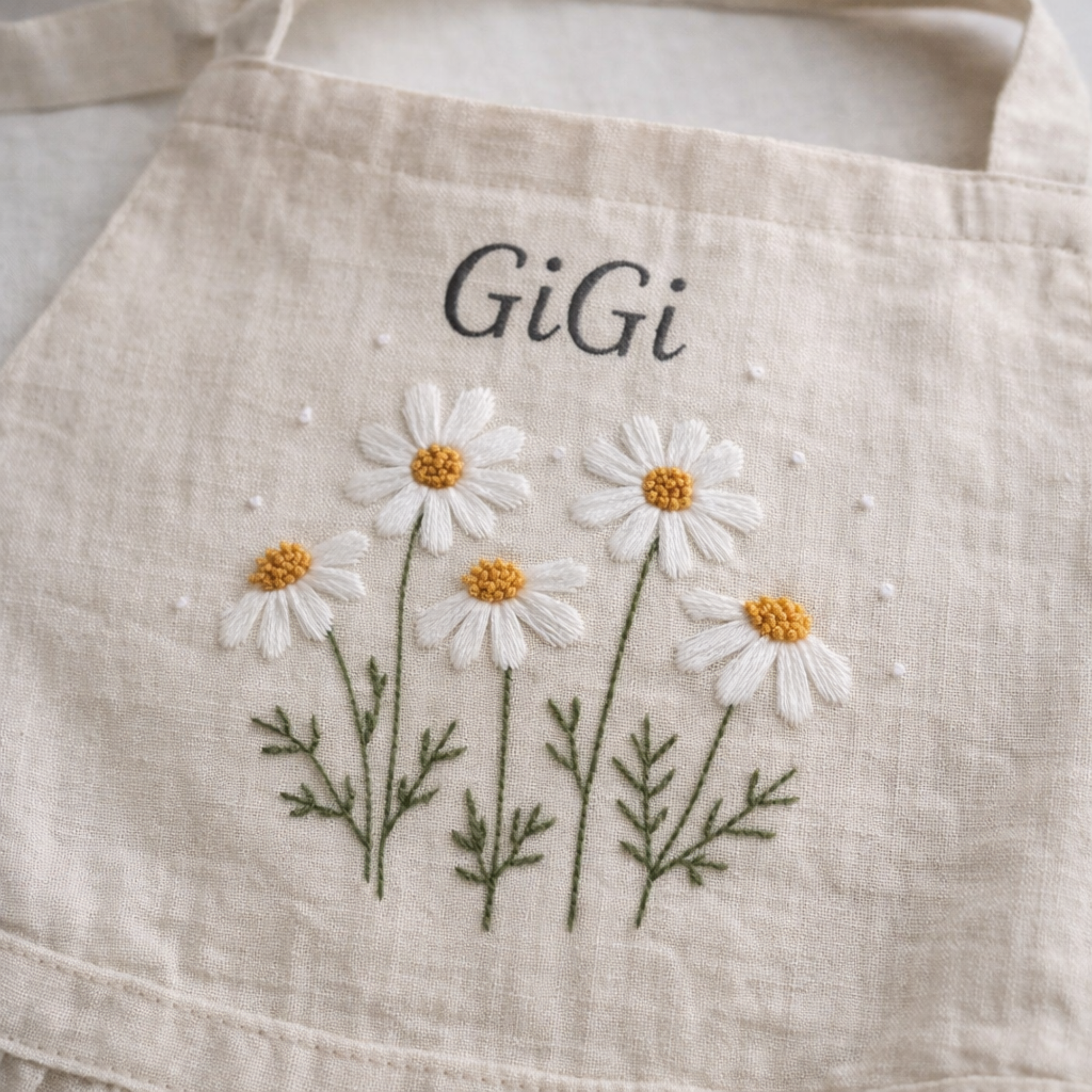 Personalized hand embroidered linen apron featuring white daisy flowers, botanical foliage, custom name embroidery, and floral pocket details.