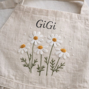 Personalized hand embroidered linen apron featuring white daisy flowers, botanical foliage, custom name embroidery, and floral pocket details.