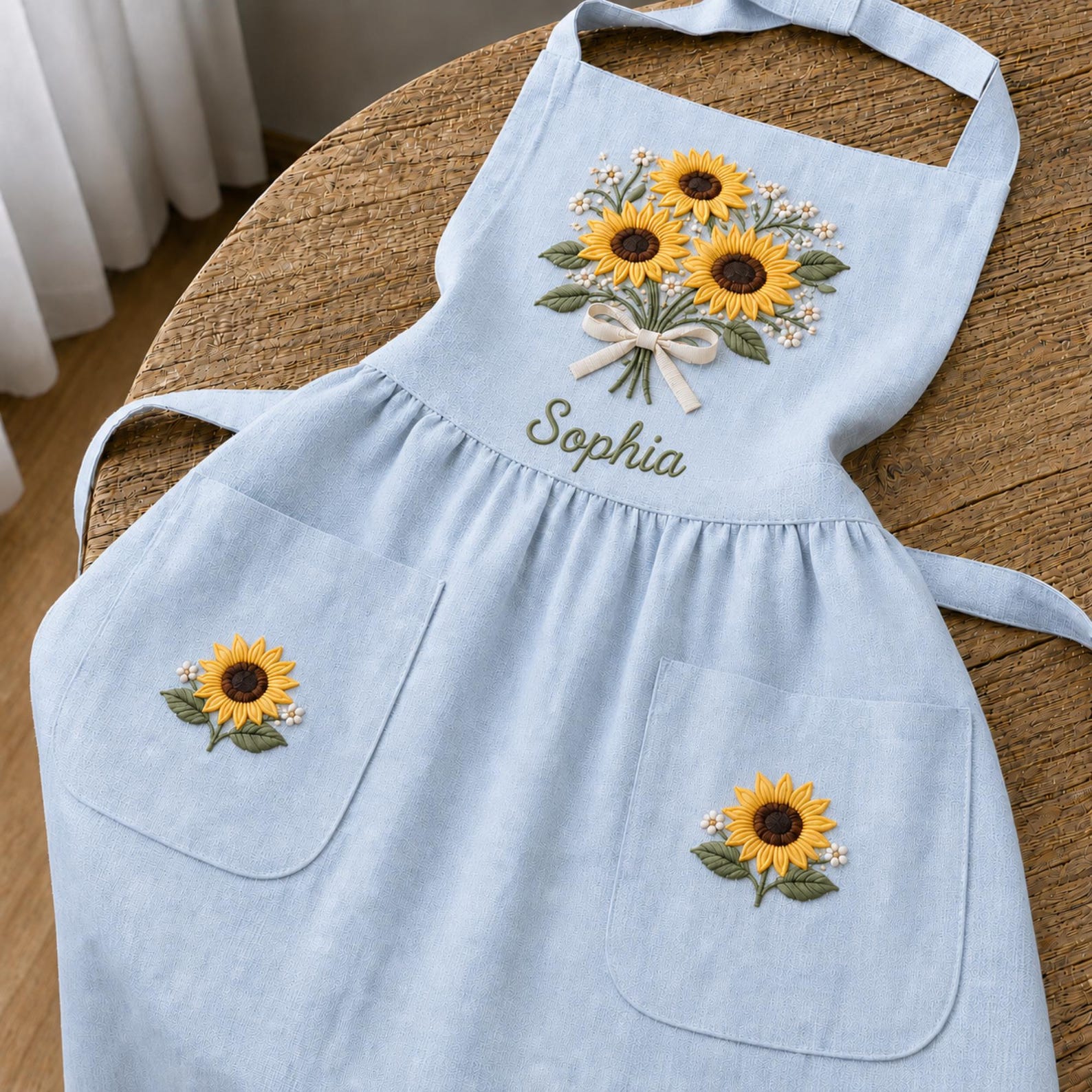 Personalized hand embroidered linen apron featuring a sunflower bouquet, white floral accents, ribbon detail, and custom name embroidery.