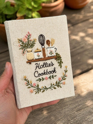Hand embroidered recipe journal featuring floral kitchen shelf with vintage jars whisk baking utensils and botanical wreath on linen cover