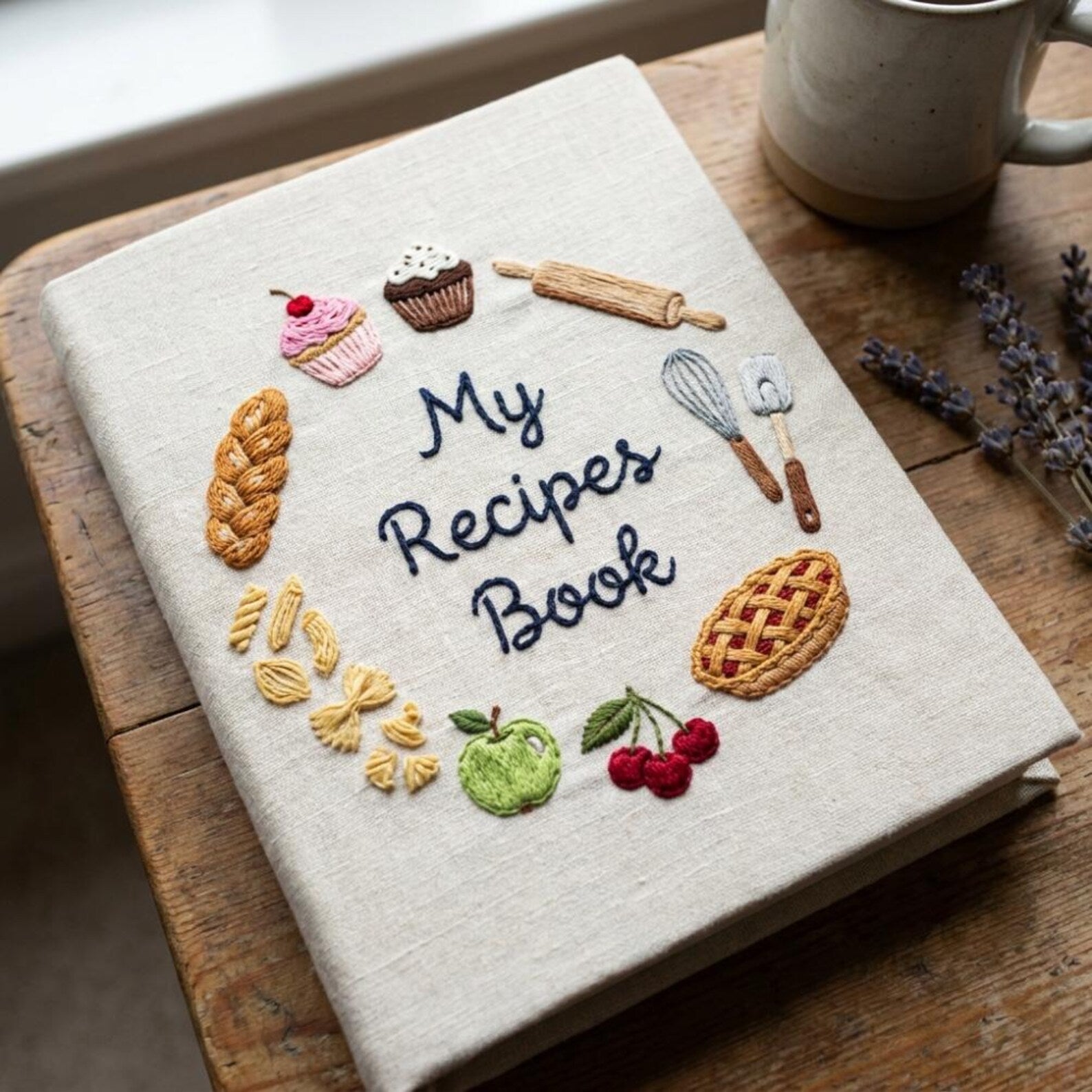 Hand embroidered recipe book featuring cupcakes, pie, artisan bread, pasta, fruit and baking tools on linen cover.
