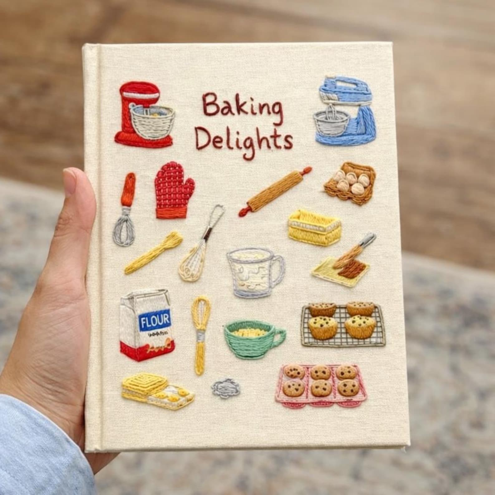 Hand embroidered recipe book featuring vintage baking studio essentials including stand mixers, rolling pins, flour, eggs, whisks, muffins and cookies on a linen cover.