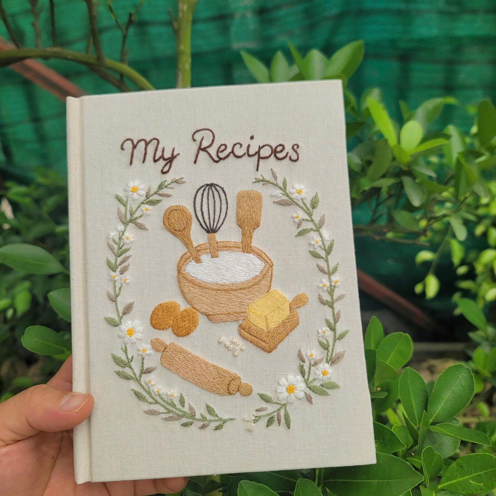 Hand embroidered recipe book featuring mixing bowl flour eggs butter rolling pin whisk and daisy floral wreath on linen cover