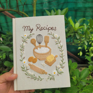 Hand embroidered recipe book featuring mixing bowl flour eggs butter rolling pin whisk and daisy floral wreath on linen cover