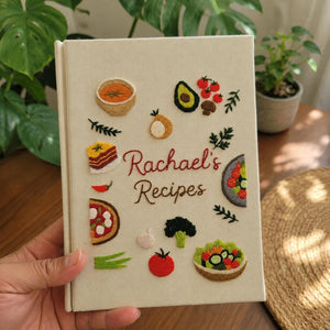 Hand embroidered recipe book featuring vegetables salads pizza lasagna soups herbs and family meal inspired food illustrations on linen cover
