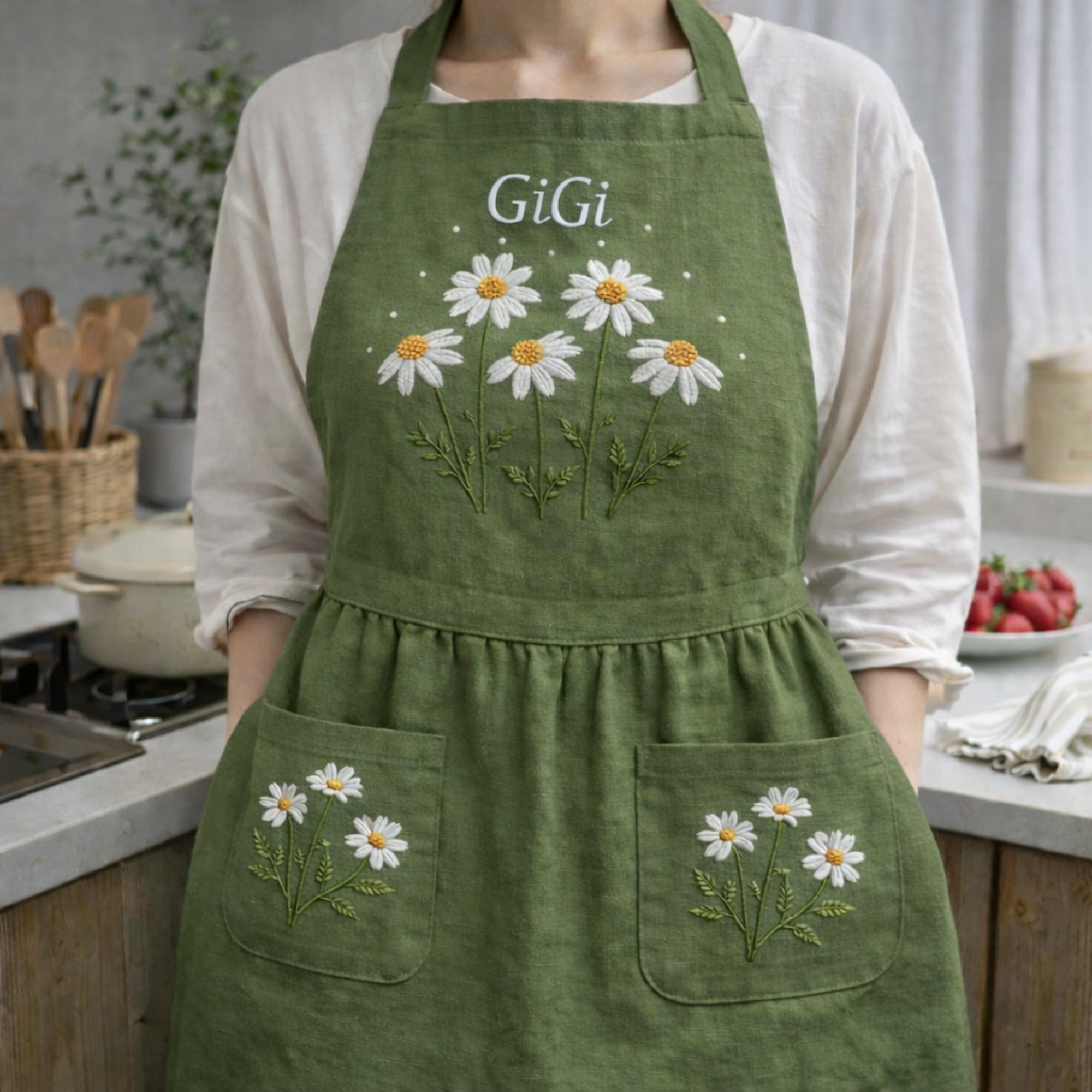 Personalized hand embroidered linen apron featuring white daisy flowers, botanical foliage, custom name embroidery, and floral pocket details.