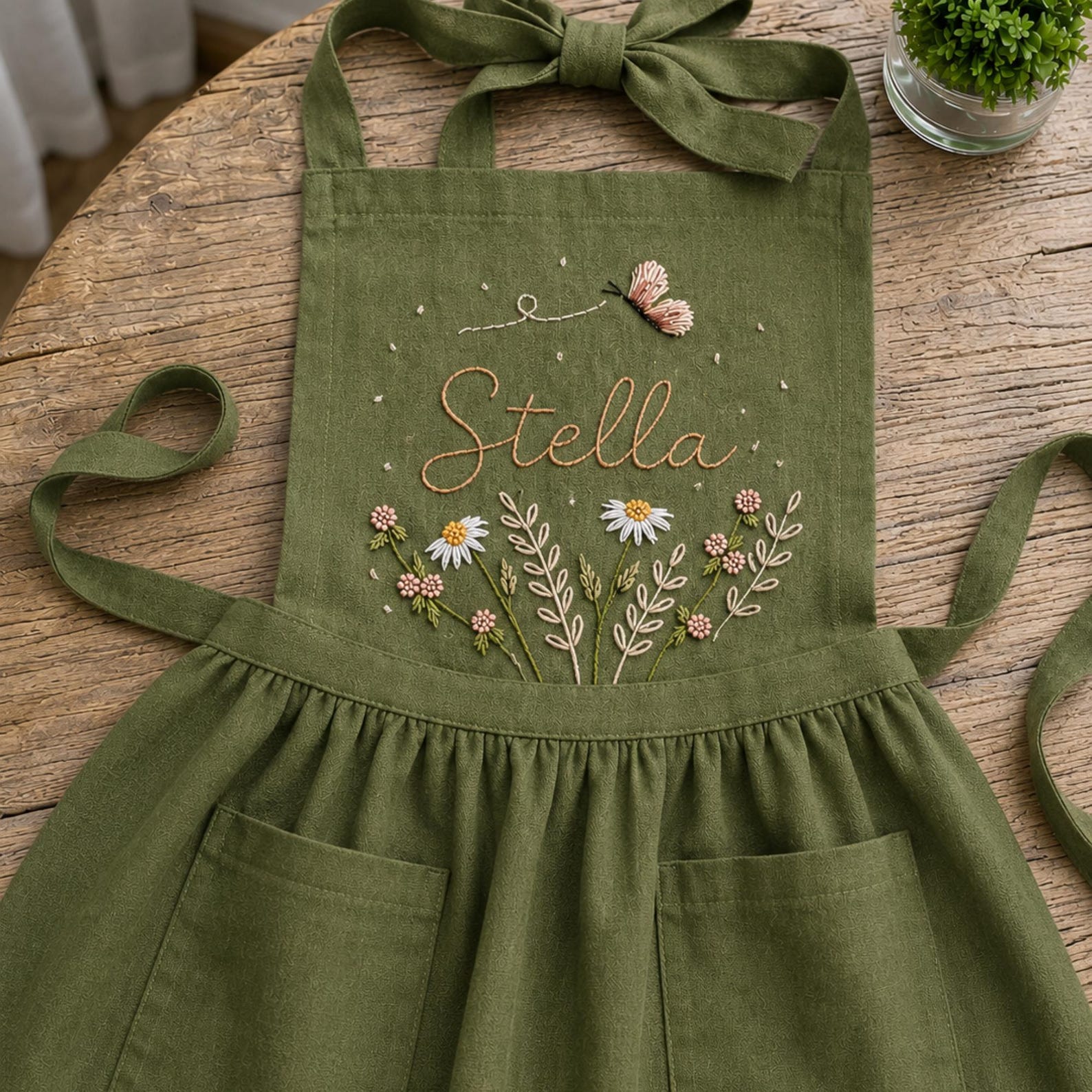 Personalized hand embroidered linen apron featuring wildflowers, daisies, botanical foliage, butterfly embroidery, and custom name embroidery on green linen fabric.