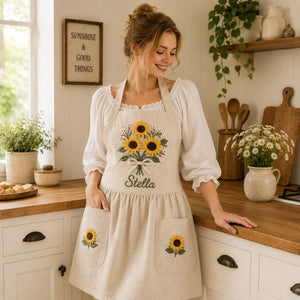 Personalized hand embroidered linen apron featuring a sunflower bouquet, white floral accents, ribbon detail, and custom name embroidery.