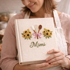 Personalized hand embroidered recipe book featuring a whisk, spatula, wooden spoon tied with a pink ribbon and surrounded by white daisies on a linen cover.